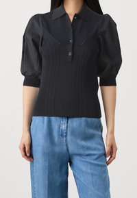 Schwarzes Strick-Poloshirt mit tailliertem Oberkörper, Puffärmeln und fünf Knöpfen. Strukturierte vertikale Linien verlaufen über die Vorderseite. Kombiniert mit hellblauem Denim.