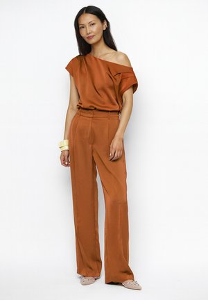 Vrouw draagt roestkleurige een-schouder blouse en hooggetailleerde wijde broek, beige versierde platte schoenen en crèmekleurige armbanden, staand tegen een witte achtergrond.