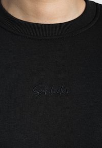 Sweatshirt en coton noir avec un col rond côtelé. Présente une broderie subtile "Smiledex" en fil foncé sur le devant. Texture lisse.