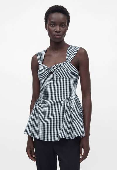 Γυναίκα που φοράει ασπρόμαυρο αμάνικο τοπ με καρό ύφασμα, φαρδιές τιράντες και πεπλούμ στο τελείωμα, σε συνδυασμό με μαύρο παντελόνι, στέκεται μπροστά σε λευκό φόντο.