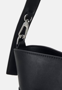 Gros plan d'une bandoulière de sac en cuir noir fixée avec un fermoir en métal argenté et un anneau au bord supérieur ouvert du sac.