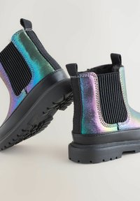 Una coppia di stivali Chelsea iridescenti con pannelli laterali elastici neri, linguette di pull e suole in gomma spesse posizionate su una superficie chiara.