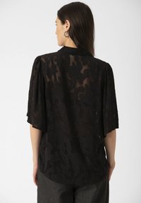 Vrouw met lang, donker haar die zilveren oorringen draagt en een sheer zwarte bloemenkant blouse met ellebooglange mouwen, gezien van achteren.