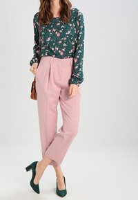 Groene blouse met bloemenpatroon, lichtroze hoge taillebroek, en groene hakken. Bruin schoudertas. Glad materiaal, nauwsluitende pasvorm.