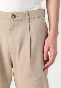 Beige színű, texturált anyagból készült nadrág, elülső redővel és gombos derékzárral. Egy kéz látható, a hátsó varrás mellett pihen.