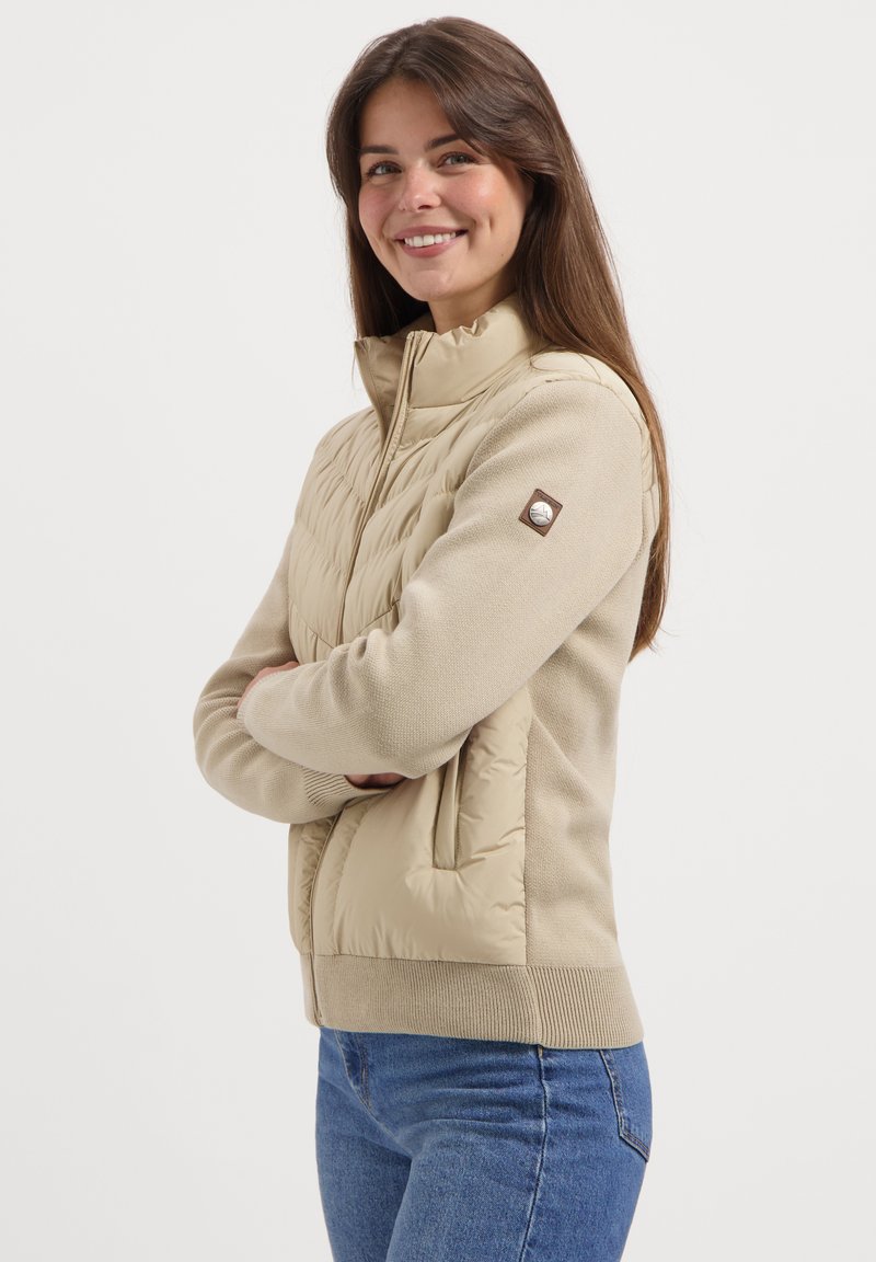 Vrouw met een glimlach en met gekruiste armen, gekleed in een beige gewatteerde jas met gebreide mouwen en een spijkerbroek, tegen een plain witte achtergrond.
