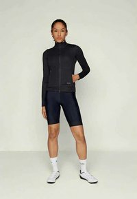 Frau in schwarzer Langarm-Reißverschlussjacke, schwarzen Radshorts, weißen Socken und weißen Radsportschuhen, die mit einer Hand in der Jackentasche steht.