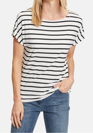 Kobieta w białej koszuli z krótkim rękawem i czarnymi poziomymi paskami oraz niebieskich dżinsach, z jedną ręką w kieszeni, na jednolitym tle.