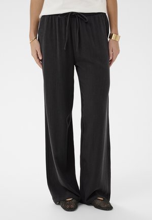 Nő laza fekete, húzózsinóros nadrágban, fekete szövött lapos cipőben, arany karkötővel és gyűrűkkel, egyszínű háttér előtt állva.