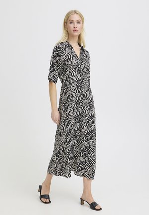 Heledate juustega naine, kes kannab musta-valge loomamustriga poolpikalt kleiti, millel on poolsed varrukad, ning mustade avatud varbaga plokkkontsaga sandaale, seisab valgel taustal.