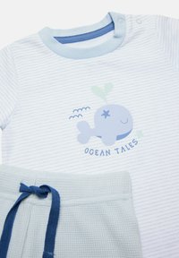 Camisa a rayas azul claro con gráfico de ballena y texto "Cuentos del Océano", combinada con pantalones cortos texturizados azul claro que presentan un cordón azul.
