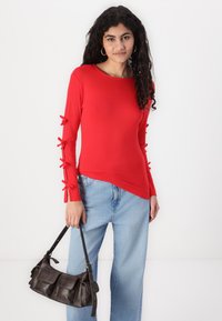 Rød langermet topp med sløyfer langs ermene, kombinert med lyseblå vide jeans og en mørkebrun veske. Glatt tekstur.