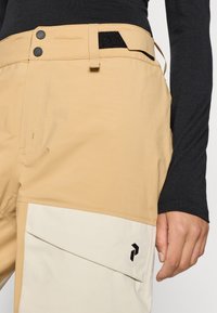 Beige broek met een gestructureerde afwerking, voorzien van zwarte hardware bij de taille, een steekzak in een lichtere tint en een logo dicht bij de zak.