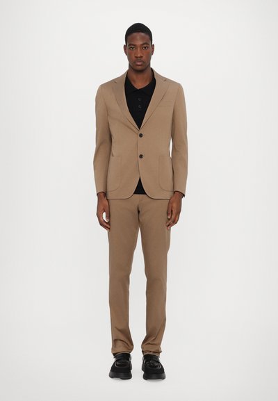 Costume beige avec un blazer à un seul boutonnage, un pantalon assorti et un polo noir en dessous. Tissu lisse, design élégant, coupe ajustée, porté avec des chaussures noires.