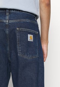 Pogled od blizu na osobu koja nosi tamnoplave traperice s Carhartt logom na zadnjem džepu i svijetlu majicu uvučenu unutra.