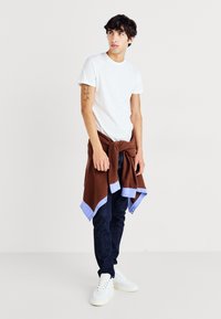 White t-shirt, brown knit sweater tied around waist with blue accents, dark jeans, and white sneakers. Casual outfit with layered textures.