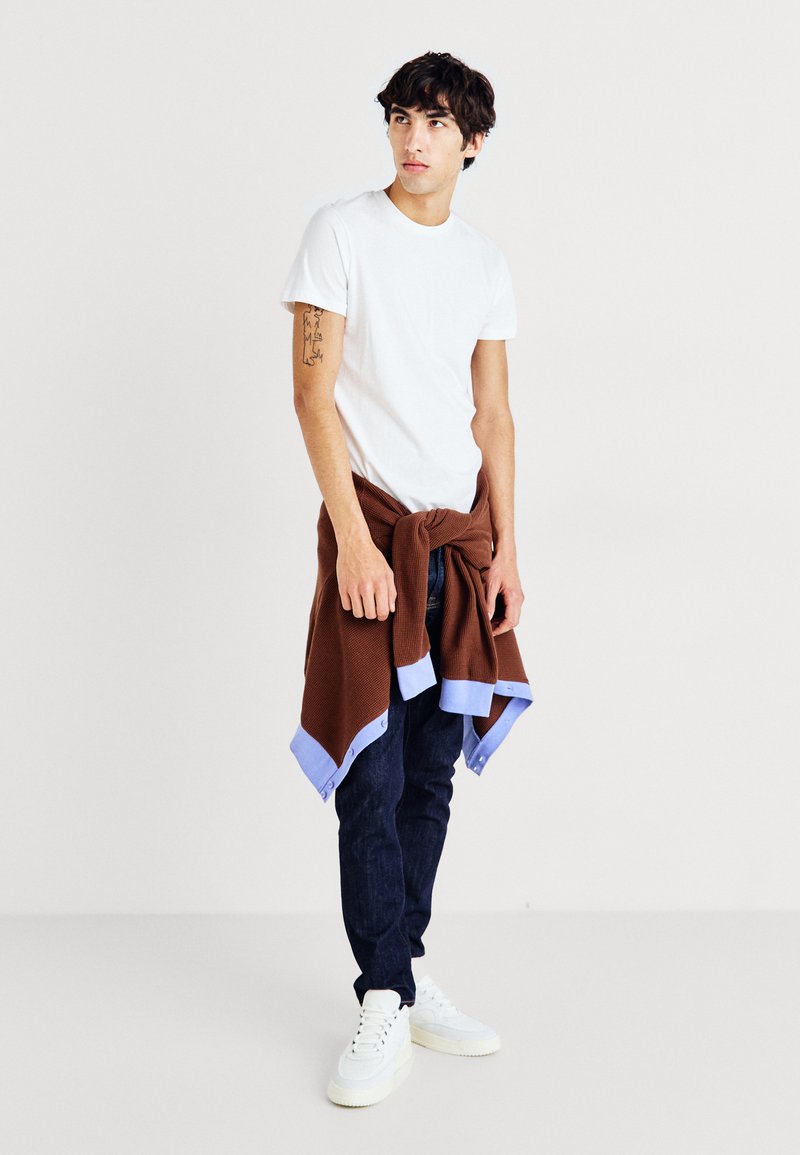 White t-shirt, brown knit sweater tied around waist with blue accents, dark jeans, and white sneakers. Casual outfit with layered textures.