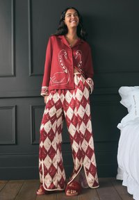 Roter, gemusterter Schlafanzug mit Schwanmotiven, langen Ärmeln und weit geschnittenen Hosen mit geometrischem Elfenbeinmuster. Flauschige rote Hausschuhe runden das Outfit ab.