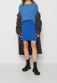 Blauer Strickpullover über einem leuchtend blauen Rock, ergänzt durch einen langen grauen Mantel und schwarze Knöchelschuhen. Glatte Stoffe, moderne Silhouette.
