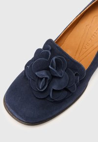 Navy suede slip-on shoe featuring a large, layered flower detail on the front, with a rounded toe and smooth inner sole.