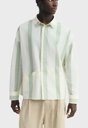 Mladić u širokoj bijeloj košulji s gumbima s uskim vertikalnim blijedo zelenim prugama i bež hlačama, gleda u stranu.