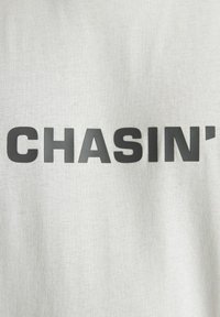 T-shirt in cotone grigio chiaro con grande testo nero "CHASIN'" sul petto, con una texture morbida e vestibilità regolare.