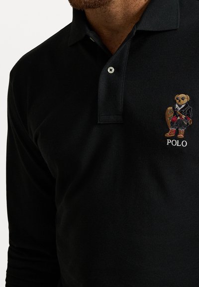 Crna polo majica s ovratnikom, izrađena od pamuka. Sadrži izvezenu medvjedu figuru u jakni, s natpisom "POLO" ispod.