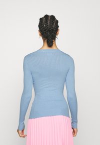 Kobieta z warkoczami, ubrana w niebieski sweter z prążkami i różową plisowaną spódnicę, stoi tyłem na tle jednolitego białego tła.