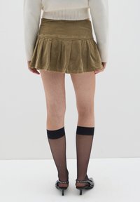 Falda mini de pana en verde olivo con pliegues, combinada con medias negras transparentes y zapatos de tacón negros con acentos plateados.