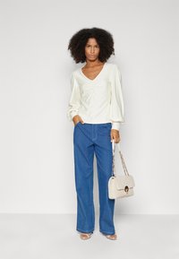 Cream V-neck top with gathered detailing, wide blue jeans, and a beige textured handbag with chain strap. Clear background.