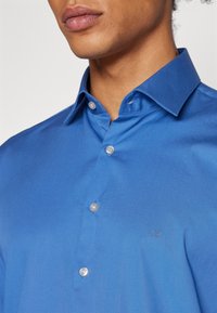 Camicia in cotone blu con colletto classico, bottoni in plastica e un piccolo logo ricamato sul lato sinistro del petto. Texture liscia.