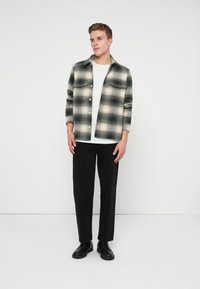 Camisa a cuadros verde y crema con botones frontales y dos bolsillos en el pecho, llevada sobre una camiseta blanca y combinada con pantalones negros de pierna recta y zapatos.