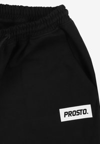 Czarne dresy z elastycznym pasem, z białą prostokątną naszywką z logo "PROSTO." na dolnej prawej stronie. Gładka tekstura.