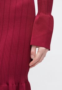 Dunkelrotes, geripptes Strickkleid mit langen Ärmeln und breiten Manschetten. Der Stoff zeigt vertikale Muster und einen ausgestellten Saum an der Unterseite.