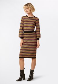 Abito a fantasia con un design ondulato in marrone, rosa, giallo e nero. Maniche lunghe, silhouette aderente e taglio al ginocchio. Stivali neri.