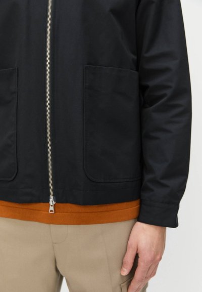 Sort zip-up jakke med to forlommer og en orange sweater underneath. Stoffet ser glat ud med en mat finish.
