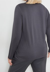 Maglietta a maniche lunghe di un grigio scuro con texture liscia. Presenta un collo rotondo e un orlo leggermente curvo. Abbinata a pantaloni scuri coordinati.