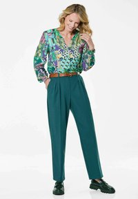 Multikleurige blouse met groene, roze en blauwe patronen; lange mouwen; V-hals. Wijde teal broek; bruine riem; glanzende donkergroene schoenen.