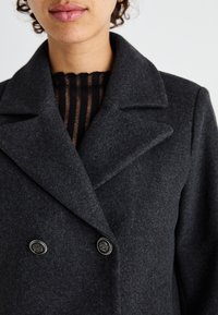 Cappotto in lana grigio scuro a doppio petto con colletto a punta, caratterizzato da bottoni di tonalità argento e una camicia interna strutturata con strisce verticali.