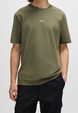 Hombre con camiseta de manga corta de color verde oliva con el logo "BOSS" centrado en el pecho y pantalones oscuros, mano en el bolsillo.
