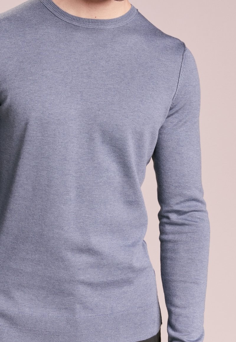 Homme portant un pull ajusté à manches longues en maille bleu clair sur un fond rose doux, visible du cou aux hanches.