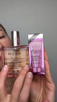 Personne tenant une bouteille d'huile sèche NUXE et un coffret cadeau shampooing cheveux NUXE sur un fond gris uni.