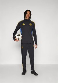 Czarny dres z żółtymi i niebieskimi akcentami, z wyszywanym logo. Osoba trzyma czarno-białą piłkę do piłki nożnej.