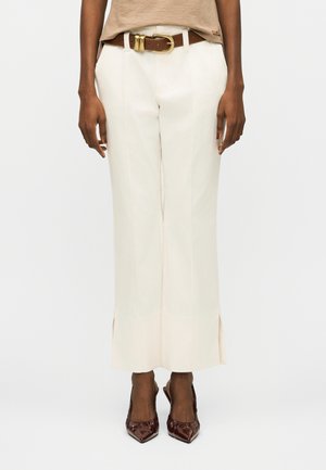 Άτομο που φοράει κρεμ παντελόνι με ψηλή μέση και φαρδιά πόδια, με ραφές στο μπροστινό μέρος, καφέ ζώνη με χρυσή αγκράφα και καφέ μυτερά τακούνια.