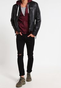 Zwarte leren jas met doorgestikte schouderdetails, bordeauxrode capuchontrui met rits, grijs T-shirt, zwarte gescheurde spijkerbroek en olijfgroene sportschoenen.