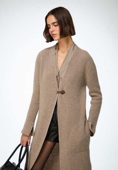 Femeie purtând un cardigan lung bej din tricot peste un tricou taupe și pantaloni scurți negri, ținând o geantă neagră, pe un fundal simplu și deschis la culoare.