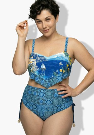 Frau mit kurzen dunklen Haaren, die einen blau gemusterten High-Waist-Bikini mit Segelboot- und Küstenlandschaftsaufdruck am Oberteil trägt.