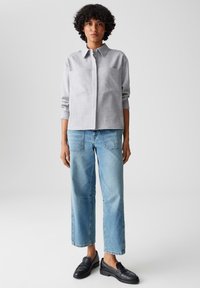 Camicia button-up grigia chiara con colletto, abbinata a jeans larghi blu chiaro e mocassini neri. La camicia presenta una tasca frontale.