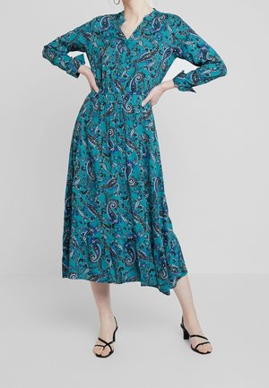 Mujer con vestido midi de manga larga color verde azulado con patrones de cachemira azules y sandalias negras de tacón, de pie con las manos en las caderas frente a un fondo liso.