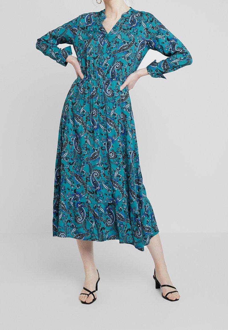 Femme portant une robe midi à manches longues bleu sarcelle avec des motifs cachemire bleus et des sandales noires à talons, debout les mains sur les hanches devant un fond uni.
